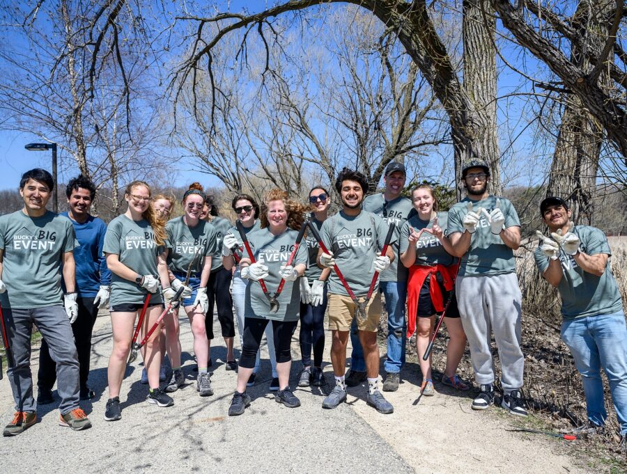 A crowd of people wearing matching T-shirts crowd together for a group photo outdoors in early spring under a large cottonwood tree. They are holding pruning sheers and smiling to the camera.