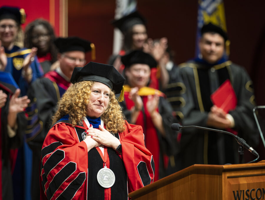 Jennifer Mnookin stands on stage with a crowd of dignataries, all wearing academic regalia. Mnookin folds her hands on her heart in a gesture of gratitude as she makes an address from a wooden podium with the word