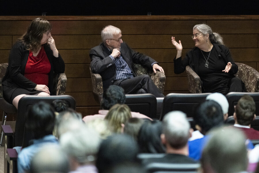 Three people sit in chairs holding a panel conversation before a seated audience.