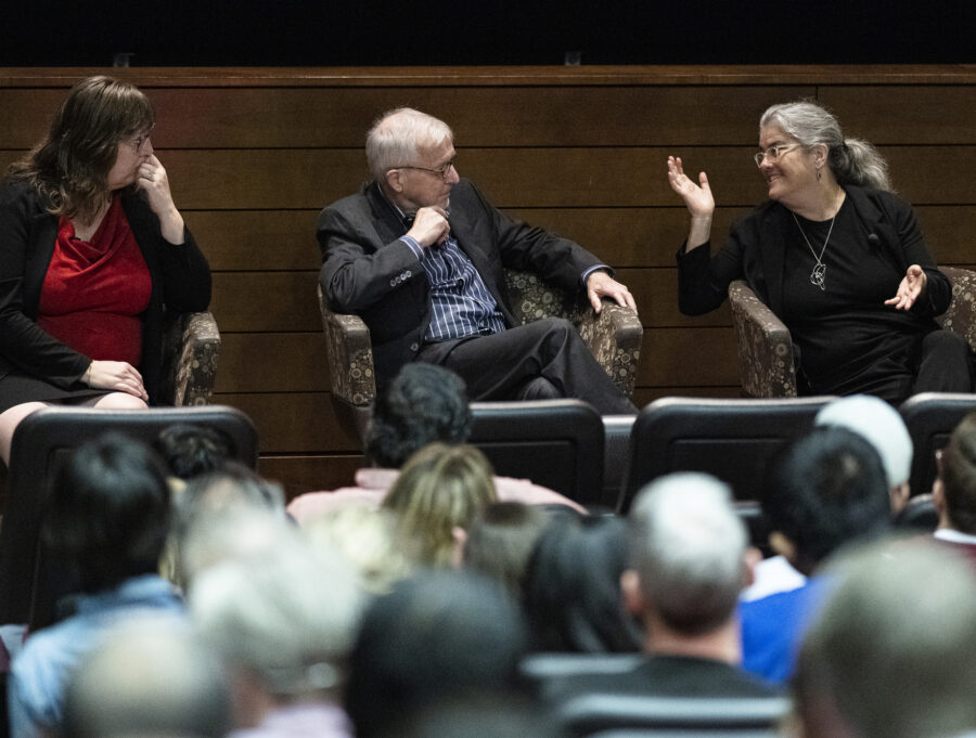 Three people sit in chairs holding a panel conversation before a seated audience.