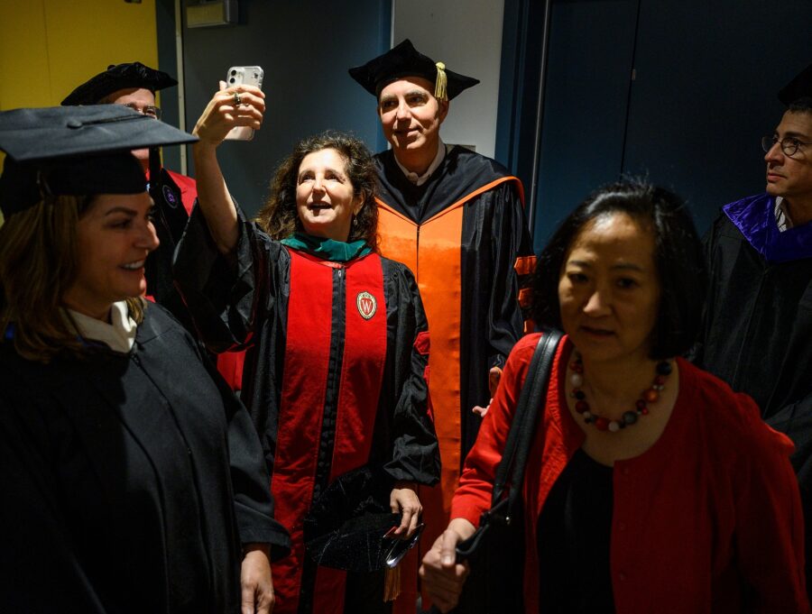 A tight shot of six people standing in a corridor wearing academic robes and hats. A woman at center raises her phone to take a selfie with another member of the group.