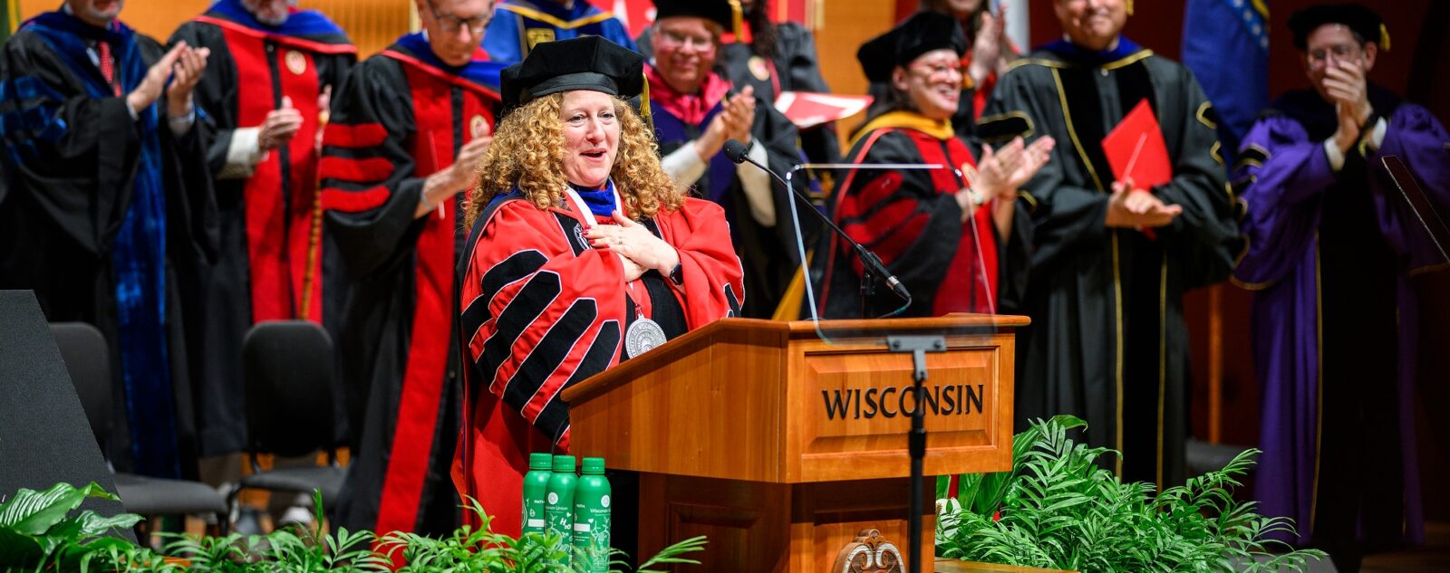 Jennifer Mnookin stands on stage with a crowd of dignitaries, all wearing academic regalia. Mnookin folds her hands on her heart in a gesture of gratitude as she makes an address from a wooden podium with the word 