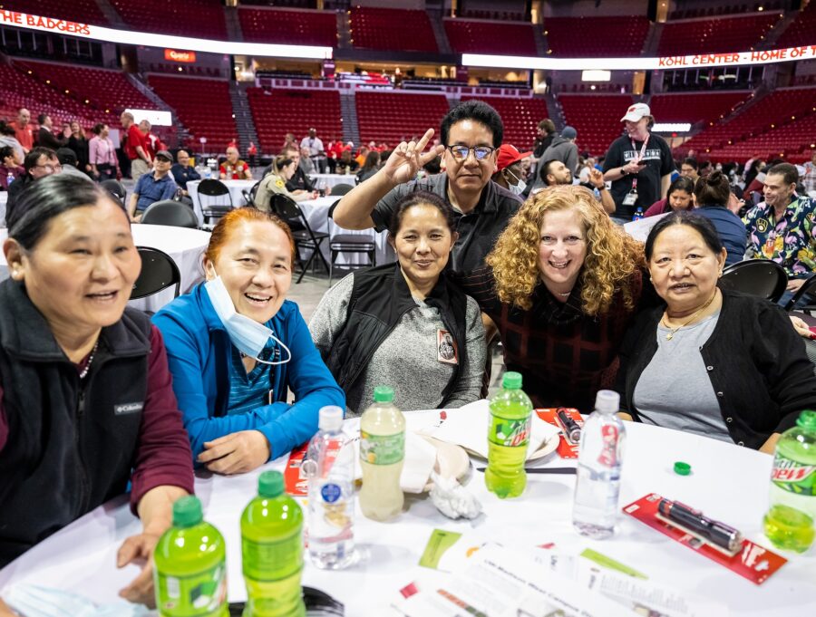 Jennifer Mnookin bends down to talk to people seated at a banquet table, posing Jennifer Mnookin leans down to take a group photo with four people seated at a banquet table on the floor of the Kohl Center arena.