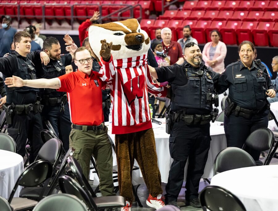On the floor of the floor of the Kohl Center arena, Bucky Badger and second- and third- shift staff, including UWPD officers in uniform, stand with arms around each other's shoulders as they smile and sing
