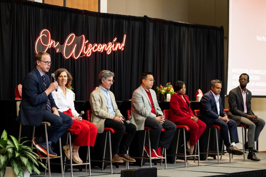Seven people sit on stools on a stage holding a panel conversation. Hanging behind them on the wall is a neon sign with the words "On, Wisconsin!"