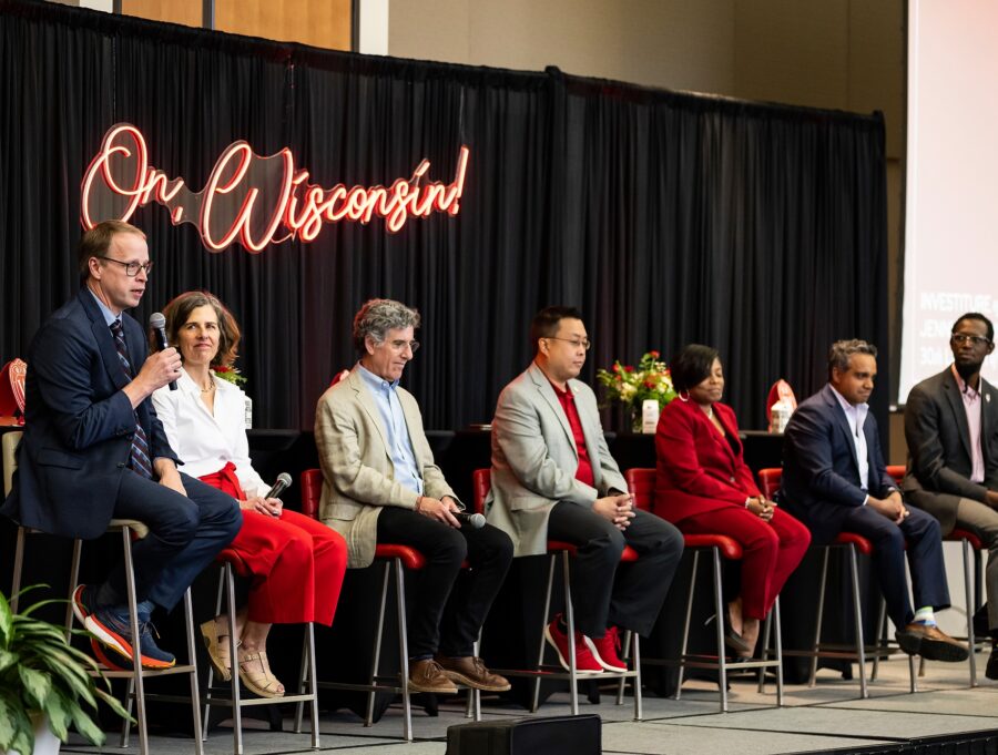 Seven people sit on stools on a stage holding a panel conversation. Hanging behind them on the wall is a neon sign with the words 